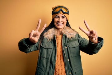 Young beautiful hispanic woman wearing ski glasses and coat for winter weather smiling with tongue out showing fingers of both hands doing victory sign. Number two.