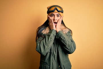 Young beautiful hispanic woman wearing ski glasses and coat for winter weather afraid and shocked, surprise and amazed expression with hands on face