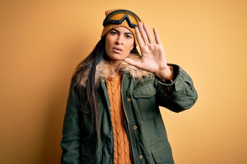 Young beautiful hispanic woman wearing ski glasses and coat for winter weather doing stop sing with palm of the hand. Warning expression with negative and serious gesture on the face.