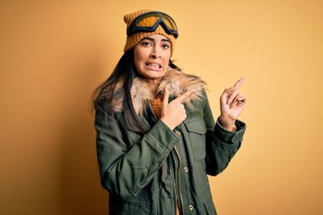 Young beautiful hispanic woman wearing ski glasses and coat for winter weather Pointing aside worried and nervous with both hands, concerned and surprised expression