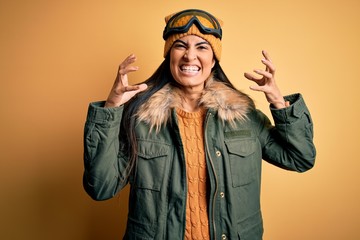 Young beautiful hispanic woman wearing ski glasses and coat for winter weather Shouting frustrated with rage, hands trying to strangle, yelling mad