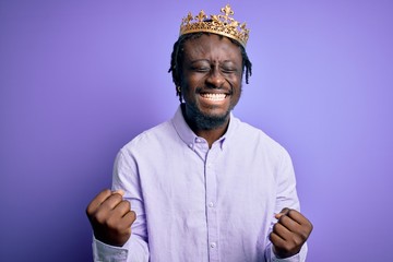 Young african american man wearing golden crown of king over isolated purple background very happy...
