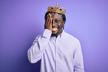 Young african american man wearing golden crown of king over isolated purple background covering one eye with hand, confident smile on face and surprise emotion.