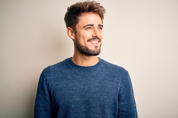Young handsome man with beard wearing casual sweater standing over white background looking away to...