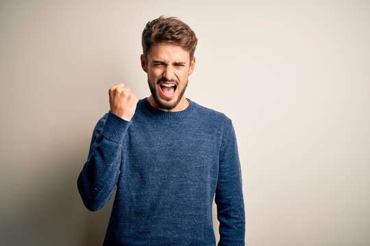 Young handsome man with beard wearing casual sweater standing over white background angry and mad raising fist frustrated and furious while shouting with anger. Rage and aggressive concept.