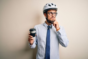 Naklejka premium Young businessman wearing glasses and bike helmet drinking cup of coffee serious face thinking about question, very confused idea