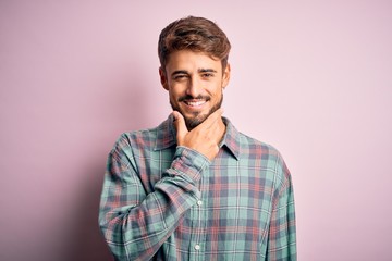 Young handsome man with beard wearing casual shirt standing over pink background looking confident at the camera smiling with crossed arms and hand raised on chin. Thinking positive.