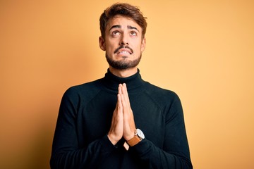 Young handsome man with beard wearing turtleneck sweater standing over yellow background begging...