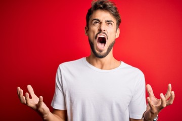 Young handsome man with beard wearing casual t-shirt standing over red background crazy and mad shouting and yelling with aggressive expression and arms raised. Frustration concept.