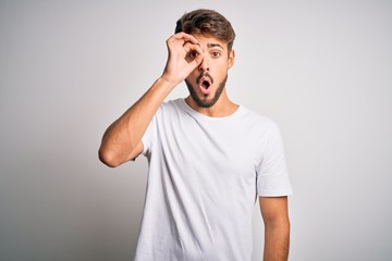 Fototapeta premium Young handsome man with beard wearing casual t-shirt standing over white background doing ok gesture shocked with surprised face, eye looking through fingers. Unbelieving expression.