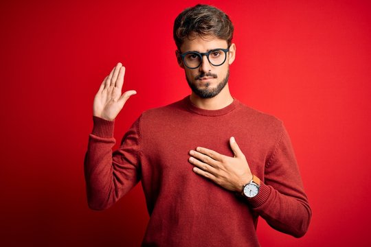 Young handsome man with beard wearing glasses and sweater standing over red background Swearing with hand on chest and open palm, making a loyalty promise oath