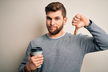 Young blond man with beard and blue eyes drinking cup of coffee over white background with angry...