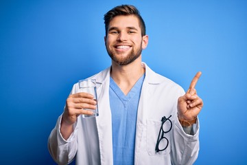 Young blond doctor man with beard and blue eyes wearing coat drinking glass of water surprised with an idea or question pointing finger with happy face, number one