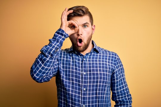 Young blond businessman with beard and blue eyes wearing shirt over yellow background doing ok gesture shocked with surprised face, eye looking through fingers. Unbelieving expression.