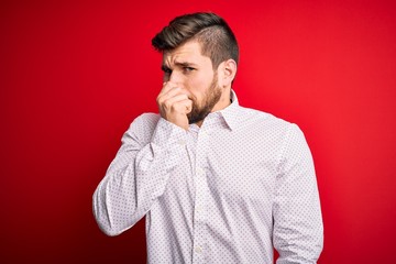Young blond businessman with beard and blue eyes wearing elegant shirt over red background smelling...