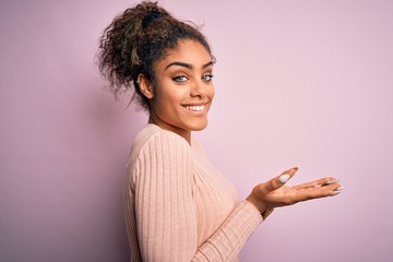 Young beautiful african american girl wearing casual sweater standing over pink background pointing aside with hands open palms showing copy space, presenting advertisement smiling excited happy