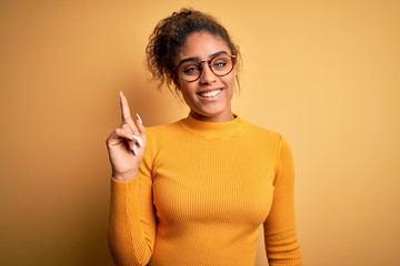 Young beautiful african american girl wearing sweater and glasses over yellow background pointing finger up with successful idea. Exited and happy. Number one.