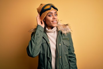 Young african american afro skier girl wearing snow sportswear and ski goggles smiling with hand over ear listening an hearing to rumor or gossip. Deafness concept.