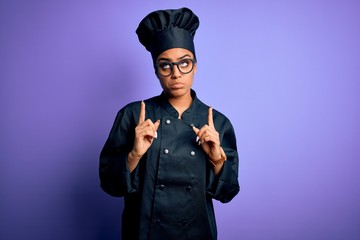Young african american chef girl wearing cooker uniform and hat over purple background Pointing up looking sad and upset, indicating direction with fingers, unhappy and depressed.