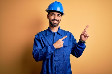 Mechanic man with beard wearing blue uniform and safety helmet over yellow background smiling and...