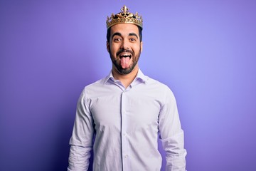 Young handsome man with beard wearing golden crown of king over purple background sticking tongue out happy with funny expression. Emotion concept.