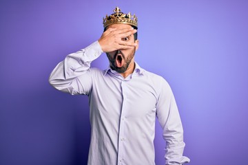 Young handsome man with beard wearing golden crown of king over purple background peeking in shock covering face and eyes with hand, looking through fingers with embarrassed expression.