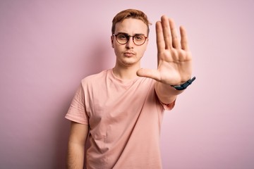 Naklejka premium Young handsome redhead man wearing casual t-shirt standing over isolated pink background doing stop sing with palm of the hand. Warning expression with negative and serious gesture on the face.
