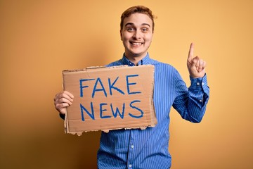 Young handsome redhead man holding banner with fake news message over yellow background surprised...