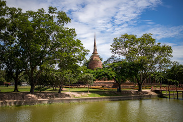 Beautiful background image of a pagoda surrounded by green trees and in front of a canal at Sukhothai Historical Park on a clear sky.