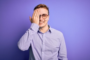 Young handsome redhead man wearing casual shirt and glasses over purple background covering one eye with hand, confident smile on face and surprise emotion.