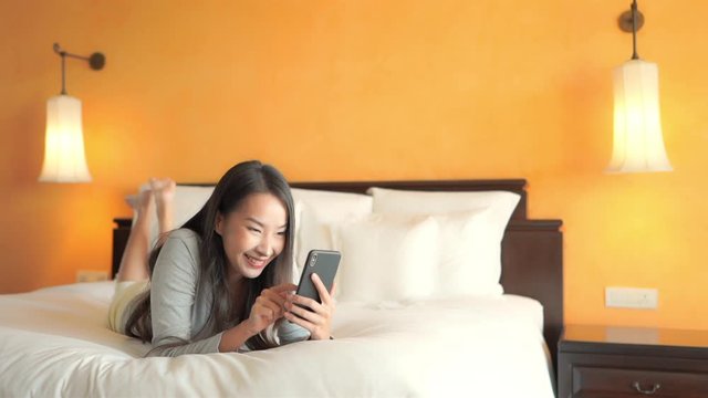  Thai woman laying on a luxury hotel bed and scrolling pictures on her phone with happy facial expression 