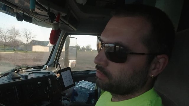 Close up of a truck driver's face while he's driving on a local highway during sunset. He keeps checking his mirrors and the road ahead is reflecting in his sunglasses.