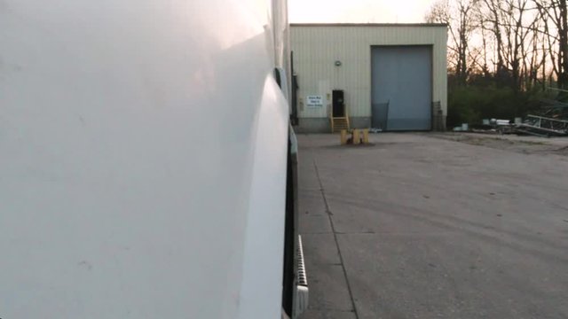 Low angle shot of a white semi truck reversing its trailer onto a dock at a warehouse during sunset. A green light is flashing on the wall of the warehouse indicating the dock is available.