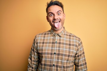Young handsome man wearing casual shirt standing over isolated yellow background sticking tongue out happy with funny expression. Emotion concept.