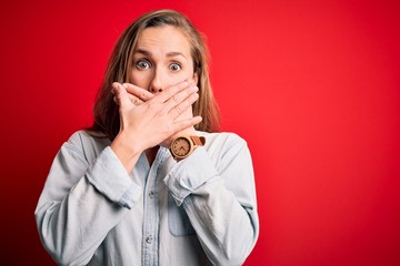 Young beautiful blonde woman wearing casual denim shirt over isolated red background shocked covering mouth with hands for mistake. Secret concept.