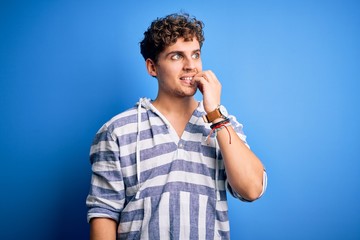 Young blond handsome man with curly hair wearing casual striped sweatshirt looking stressed and nervous with hands on mouth biting nails. Anxiety problem.