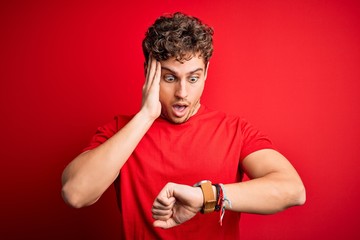 Young blond handsome man with curly hair wearing casual t-shirt over red background Looking at the watch time worried, afraid of getting late