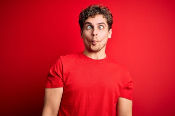 Obraz premium Young blond handsome man with curly hair wearing casual t-shirt over red background making fish face with lips, crazy and comical gesture. Funny expression.
