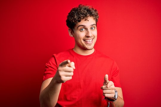 Young blond handsome man with curly hair wearing casual t-shirt over red background pointing fingers to camera with happy and funny face. Good energy and vibes.