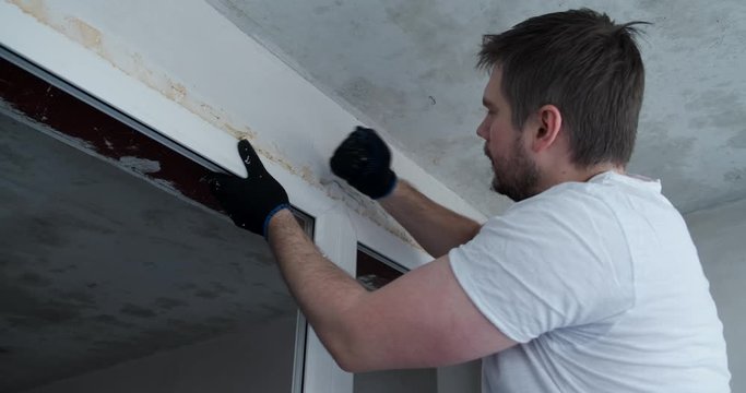 Male Builder With Knife Cuts Mounting Foam Around Perimeter Of Plastic Window Opening, Pieces Of Foam Are Flying In Different Directions, Worker Is Passionate About Work, Room Is Not Being Repaired