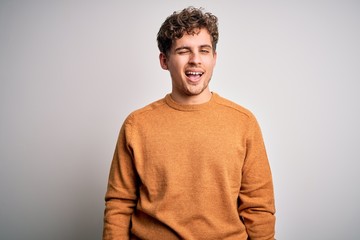 Fototapeta premium Young blond handsome man with curly hair wearing casual sweater over white background winking looking at the camera with sexy expression, cheerful and happy face.