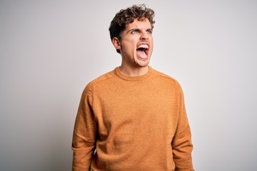 Fototapeta premium Young blond handsome man with curly hair wearing casual sweater over white background angry and mad screaming frustrated and furious, shouting with anger. Rage and aggressive concept.