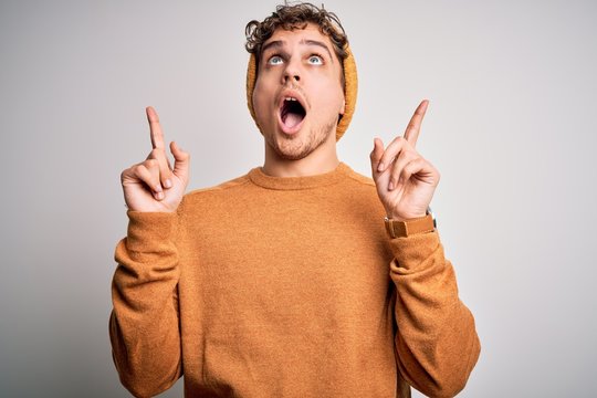 Young blond handsome man with curly hair wearing casual sweater and wool hat amazed and surprised looking up and pointing with fingers and raised arms.