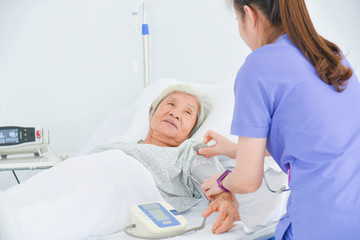 Senior asian female patient lying on bed while nurse measuring blood pressure in hospital ward.