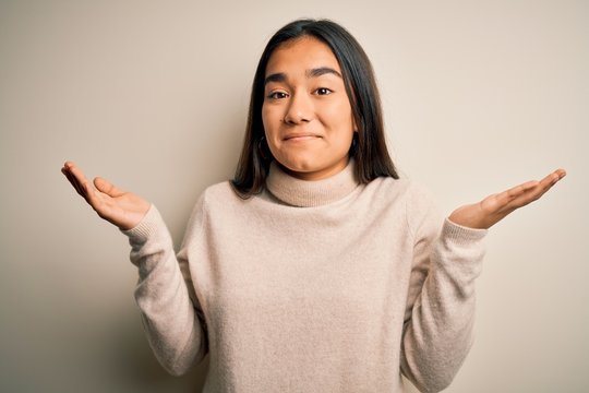 Young beautiful asian woman wearing casual turtleneck sweater over white background clueless and confused expression with arms and hands raised. Doubt concept.