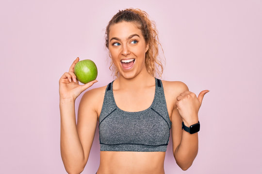 Young beautiful blonde sporty woman with blue eyes holding healthy green apple fruit pointing and showing with thumb up to the side with happy face smiling