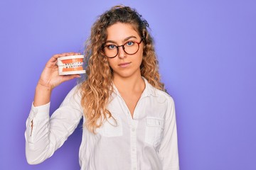 Beautiful blonde woman with blue eyes holding plastic dentrure teeth over purple background with a confident expression on smart face thinking serious