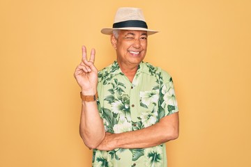 Middle age senior grey-haired man wearing summer hat and floral shirt on beach vacation smiling with happy face winking at the camera doing victory sign. Number two.
