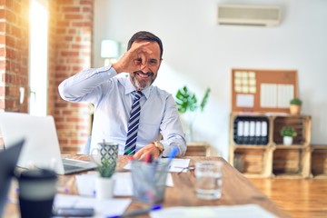 Middle age handsome businessman wearing tie sitting using laptop at the office doing ok gesture with hand smiling, eye looking through fingers with happy face.