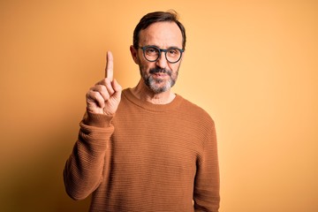 Middle age hoary man wearing brown sweater and glasses over isolated yellow background showing and pointing up with finger number one while smiling confident and happy.
